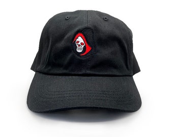 May include: A black baseball cap with an embroidered red and white skull design. The skull is wearing a red hooded cloak.