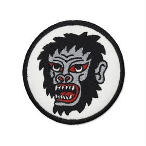 Peut inclure: Écusson brodé circulaire représentant un gorille en colère avec des yeux et des dents rouges. Le gorille a des poils noirs et un visage gris, sur fond blanc avec une bordure noire. Idéal pour personnaliser des vêtements.
