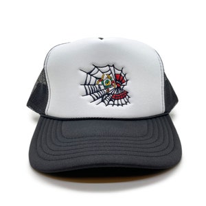 May include: A white and black trucker hat with a black mesh back. The front of the hat features an embroidered design of a spider web with a red and green eye in the centre.