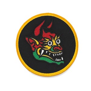 May include: Embroidered patch with a demon face design. The patch features red horns, a yellow and green face, and white teeth, set against a black background. It has a yellow border.