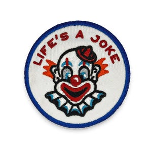 Könnte beinhalten: Runder, gestickter Aufnäher mit einem Clown-Gesicht-Design. Der Aufnäher hat einen weißen Hintergrund und einen blauen Rand. Der Clown hat eine rote Nase, Mund und Hut mit blauen und weißen Akzenten. Der Text "LIFE'S A JOKE" ist rot gestickt.