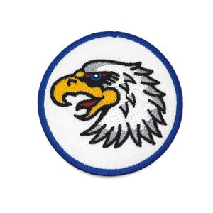 May include: Embroidered patch with an eagle's head in profile. The eagle has a yellow beak, red mouth, and blue eye, set against a white background. The design is framed by a blue border, creating a striking emblem.