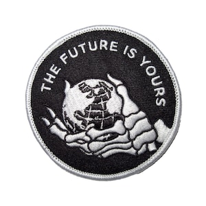 May include: A black and white embroidered patch with a skeleton hand holding a globe. The text "THE FUTURE IS YOURS" is written around the globe.