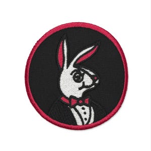 Peut inclure: Écusson brodé circulaire avec un lapin blanc portant un smoking et un nœud papillon sur fond noir. Le patch a une bordure rouge. Les oreilles du lapin ont des détails rouges.