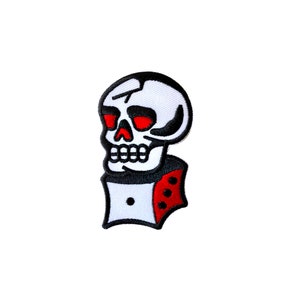 May include: White skull with red eyes and a black outline, wearing a white and red dice with black dots. The skull is facing to the right.