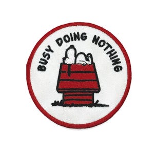Peut inclure: Écusson brodé représentant Snoopy dans sa niche rouge, avec le texte "BUSY DOING NOTHING" dans un design circulaire. L'écusson a un fond blanc avec une bordure rouge.