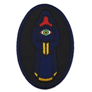 May include: Embroidered patch featuring a hooded figure with an eye symbol. The figure is in a dark blue robe with a red hood and yellow accents. The eye is green with a red outline, set against a black oval background.
