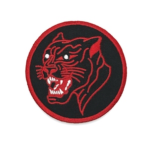 May include: Circular embroidered patch with a red panther head design on a black background. The panther has white teeth and eyes, and the patch is edged with red. Ideal for customising clothing or accessories.
