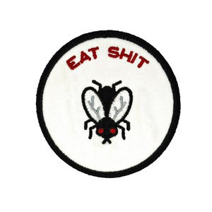 May include: Embroidered patch with a white background, black border, and the words "EAT SHIT" in red. A black fly with red eyes and gray wings is in the center. The patch is round.