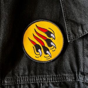 May include: A yellow circle patch with a black and white bear claw print with red accents. The patch is sewn onto a black fabric background.