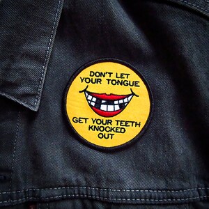 May include: Yellow embroidered patch with a smiling face and the text "Don't let your tongue get your teeth knocked out."