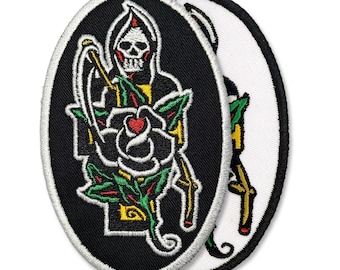 Puede incluir: Dos parches bordados de forma ovalada. El parche principal presenta una figura de la Parca con una guadaña, una rosa y una calavera. El diseño incorpora colores negro, blanco, rojo, amarillo y verde. El segundo parche es una imagen especular.