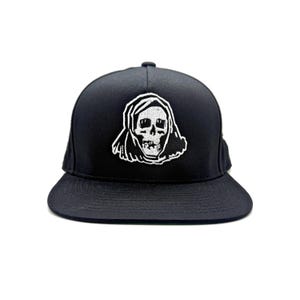 May include: Black snapback hat with a white embroidered skull design. The skull is wearing a hood. The hat has a flat brim and a structured crown. This hat is a great accessory for anyone looking for a unique and edgy style.