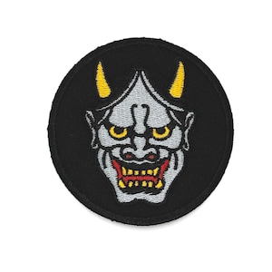May include: Round black embroidered patch featuring a stylized Japanese demon face. The demon has gray skin, yellow horns and eyes, and a red mouth with yellow teeth. The patch is a decorative item.