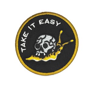May include: Embroidered patch with a black background and a yellow border. The patch features a skull-shaped snail with the words "TAKE IT EASY" in white. The snail is white and yellow.