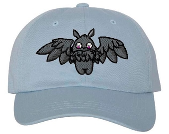 May include: A pale blue baseball cap with an embroidered cartoon bat design. The bat has grey wings and body, and pink eyes. The cap has a curved peak and a button on top.