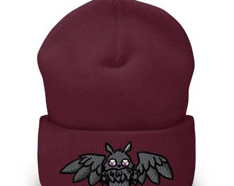 May include: A burgundy beanie hat with an embroidered cartoon bat design. The bat has large wings and purple eyes. The design is on the folded brim. The hat is made of a soft, knitted material.