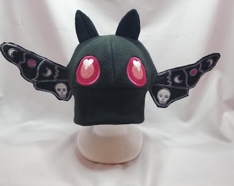 May include: Black fleece hat with bat wings and pink heart eyes. The wings have embroidered skulls and moons.