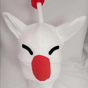 Puede incluir: Sombrero de felpa blanco con orejas grandes y puntiagudas, ojos negros curvos y una nariz roja. Un tallo blanco con un pompón rojo se extiende desde la parte superior. El sombrero está diseñado para parecerse a un personaje de videojuego.