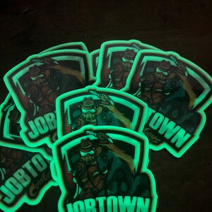 May include: A collection of glowing stickers featuring a cowboy riding a horse. The stickers have a shield-shaped border with the words "JOBTOWN Cowboys" in white and a neon green glow. The cowboy is wearing a hat and holding a rope.