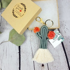Pode incluir: Um chaveiro de cacto verde e branco com pompons laranja. O chaveiro está em uma pequena caixa marrom com um adesivo de agradecimento. O adesivo diz "Obrigado por apoiar meu pequeno negócio".