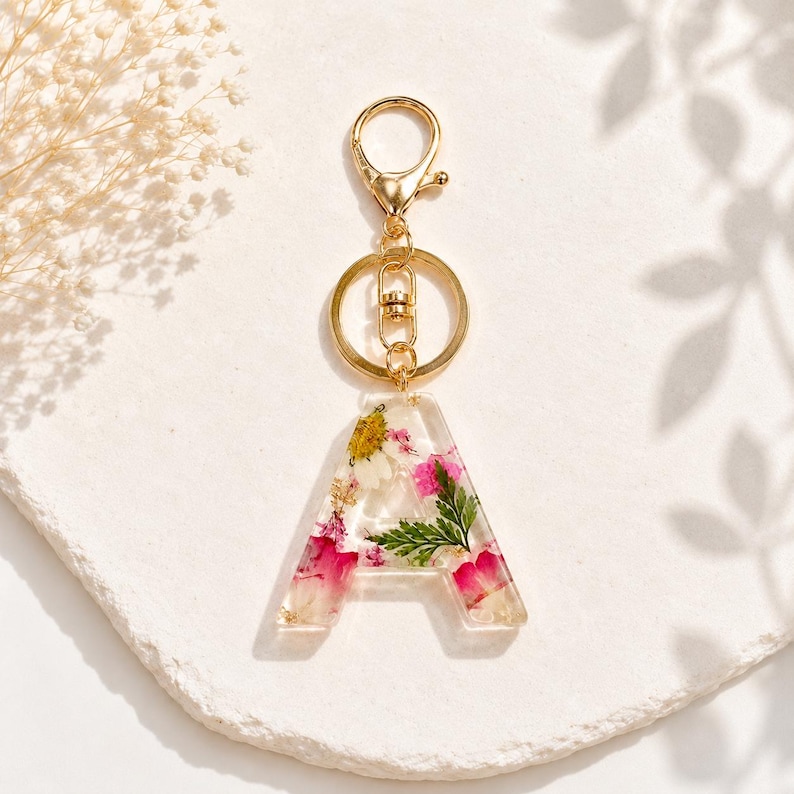 May include: A clear resin keychain in the shape of the letter "A", filled with dried pink, white, and yellow flowers, and gold flakes. The keychain has a gold-colored clasp and ring.