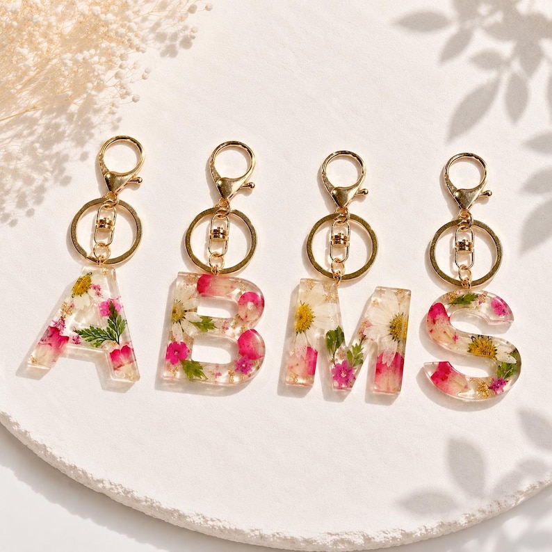 May include: Four clear resin keychains spelling out the letters A, B, M, and S. Each letter is filled with dried pink and white flowers and gold-colored hardware.
