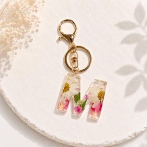 May include: A clear resin keychain with the letter "M" embedded with dried flowers in shades of white, pink, and yellow. The keychain has a gold-colored clasp and ring.