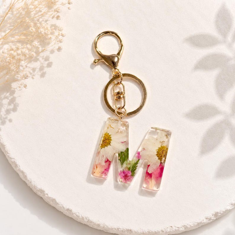 May include: A clear resin keychain with the letter "M" embedded with dried flowers in shades of white, pink, and yellow. The keychain has a gold-colored clasp and ring.