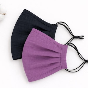 Może przedstawiać: Dwie maski na twarz, jedna czarna i jedna fioletowa, z regulowanymi czarnymi pętelkami na uszy. Maski wykonane są z teksturowanej tkaniny i leżą na białej powierzchni. Gałązka bawełny dodaje dekoracyjnego akcentu.