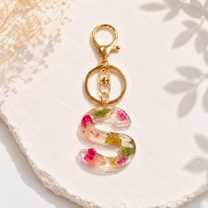 May include: A clear resin letter "S" keychain with embedded pink and yellow flowers and gold flakes. The keychain has a gold-colored clasp and ring for attaching to keys or a bag. The background is a textured white surface.