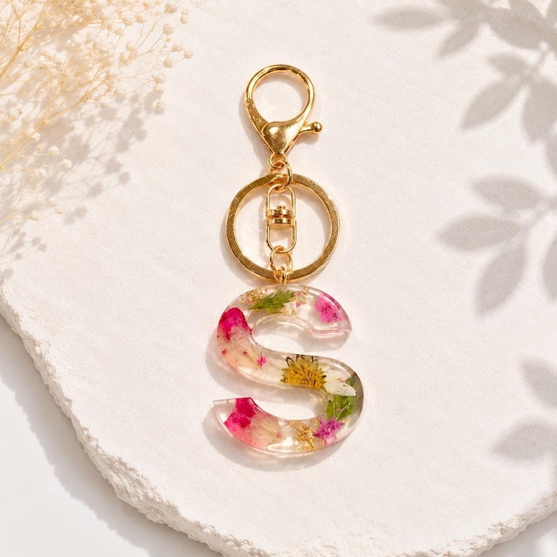 May include: A clear resin letter "S" keychain with embedded pink and yellow flowers and gold flakes. The keychain has a gold-colored clasp and ring for attaching to keys or a bag. The background is a textured white surface.
