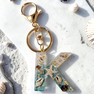 Peut inclure: Un porte-clés doré avec la lettre "K" en résine transparente. La résine est remplie de coquillages, d'une étoile de mer et de paillettes dorées. Le porte-clés est sur une surface blanche avec du sable et des coquillages.