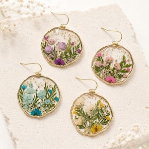 May include: Four round drop earrings with gold-toned edges, each containing pressed dried flowers in clear resin. The flowers feature varied color palettes including purple, pink, blue, and yellow, paired with green foliage and gold flakes.