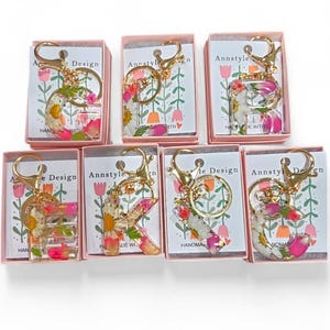 May include: A collection of floral resin letter keychains, each with a gold-colored clasp and ring. The letters are filled with dried flowers in shades of pink, white, and yellow. Each keychain is packaged in a small pink box.
