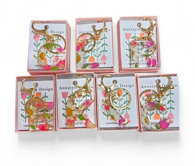May include: A collection of floral resin letter keychains, each with a gold-colored clasp and ring. The letters are filled with dried flowers in shades of pink, white, and yellow. Each keychain is packaged in a small pink box.