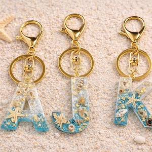 May include: Three resin letter keyrings spelling "AJK" with gold-coloured hardware. The letters are filled with blue and clear resin, gold flakes, and miniature seashells and starfish. The keyrings are on a sandy surface.