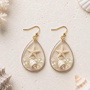 May include: A pair of teardrop-shaped earrings featuring a clear resin fill containing a small white starfish, a tiny spiral seashell, and iridescent crushed shell fragments. The earrings hang from gold-toned metal hooks with a simple wire design.