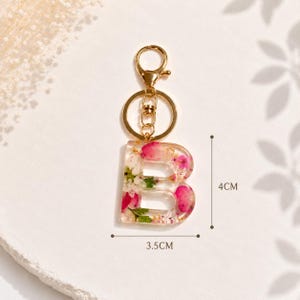 May include: A clear resin keychain in the shape of the letter "B", filled with pink and white flowers and gold flakes. The keychain is 1.4 inches wide and 1.6 inches tall, with a gold-colored metal clasp and keyring.