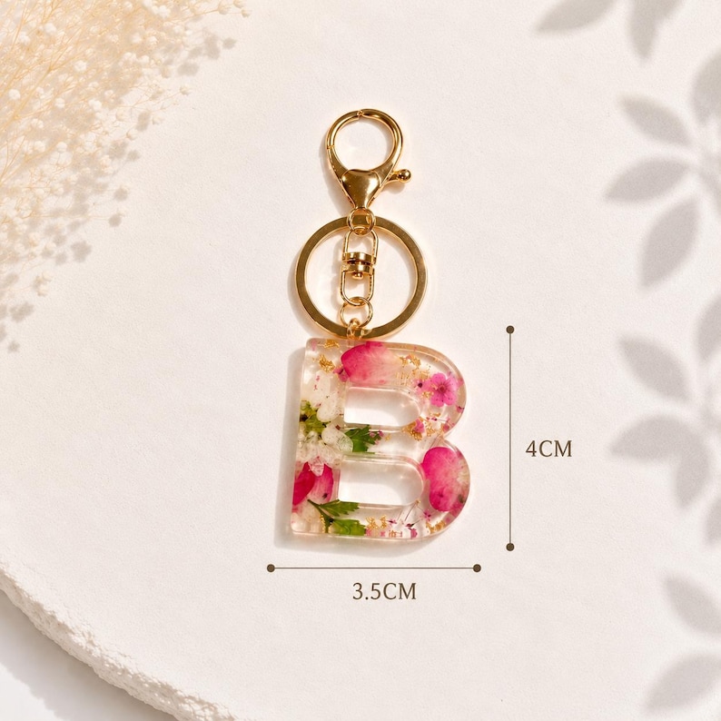 May include: A clear resin keychain in the shape of the letter "B", filled with pink and white flowers and gold flakes. The keychain is 1.4 inches wide and 1.6 inches tall, with a gold-colored metal clasp and keyring.