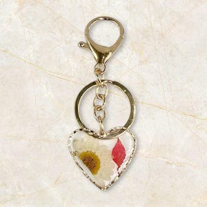 May include: Gold-toned keychain with a heart-shaped pendant. The pendant is filled with a white daisy and a red leaf, encased in clear resin. The keychain has a clasp and a ring for keys.