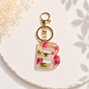 May include: A clear resin keychain in the shape of the letter "B", filled with pink and white flowers and gold flakes. The keychain has a gold-colored clasp and key ring.