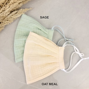 May include: Two face masks, one sage green and one oatmeal, with white elastic straps. The masks are made of a soft, breathable fabric.