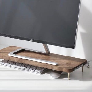 May include: A wooden monitor stand with a silver monitor on top. The stand has a built-in cable management system and gold-coloured legs. A keyboard and mouse are on the desk in front of the stand.