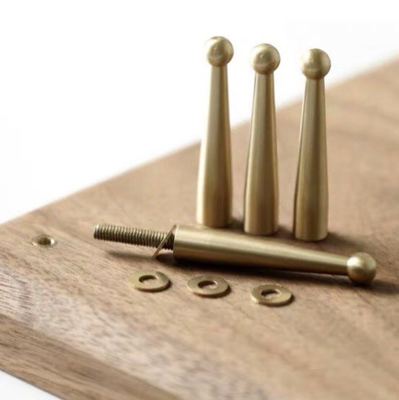May include: Four gold-toned metal furniture legs with rounded tops. The legs are shown on a wooden surface with a screw and washers.