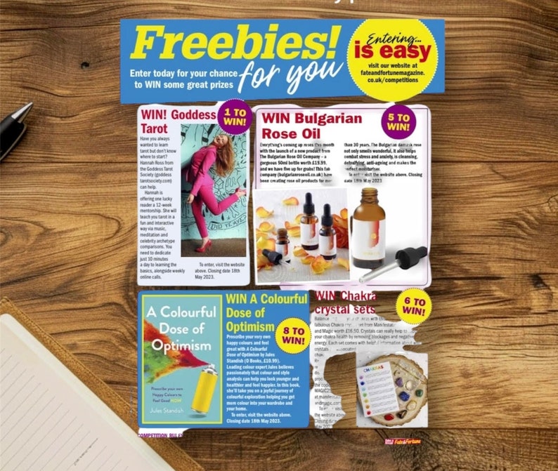 May include: A promotional flyer titled Freebies! for you featuring multiple giveaway sections including Goddess Tarot, Bulgarian Rose Oil, A Colourful Dose of Optimism, and Chakra crystal sets, with text details and product imagery on a wood grain background.