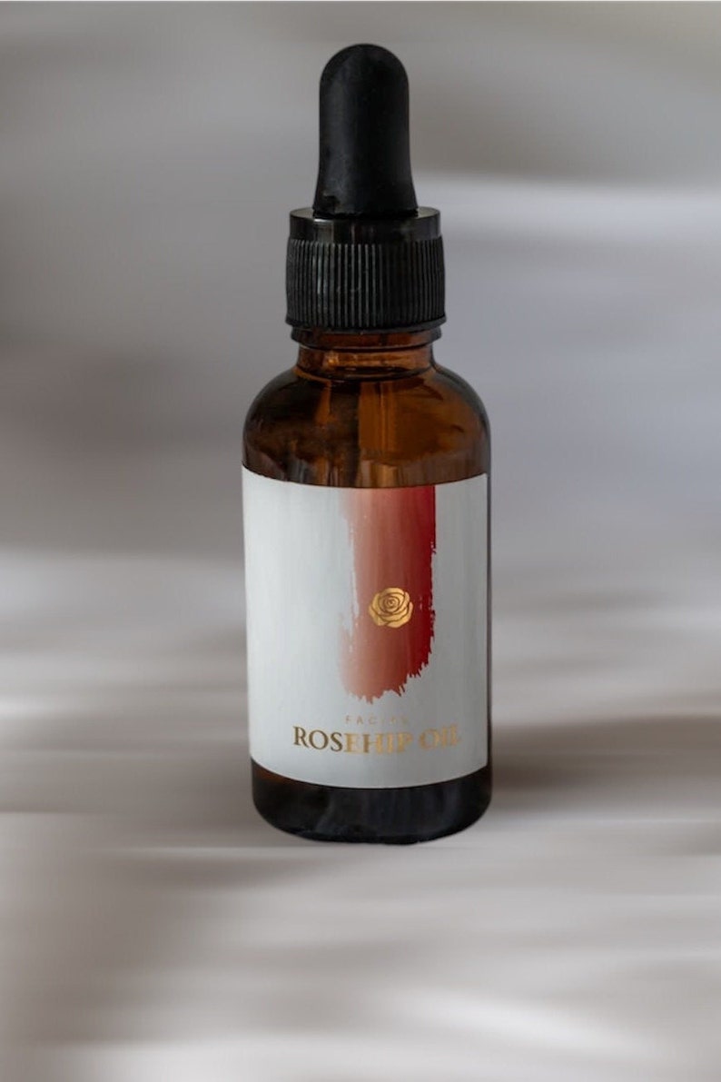 May include: An amber glass bottle with a black dropper cap, featuring a white label with a red brushstroke design, a gold rose icon, and the text "FACIAL ROSEHIP OIL" printed in gold lettering on the front of the bottle.