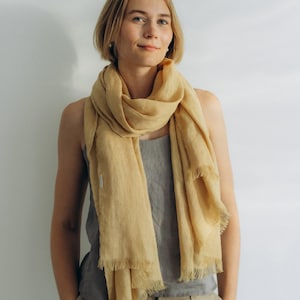 May include: A woman wearing a light brown linen scarf with frayed edges. The scarf is draped around her neck and tied in a loose knot. She is wearing a gray tank top and beige pants.