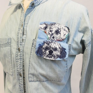 May include: Light blue denim shirt with two front pockets. The left pocket features a light blue patch with a black and white illustration of a dog. The shirt has a button-down front and long sleeves.