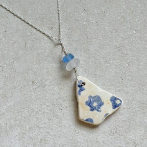 May include: A silver chain necklace featuring a pendant made from a triangular piece of white ceramic with a blue floral pattern. The pendant is attached to the chain with a wire-wrapped connection that includes one small blue bead and one clear bead.